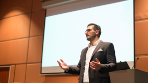 A well dressed event emcee standing in front of a projector speaking in public 