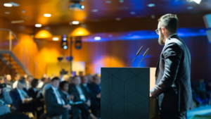 An MC standing at a podium with 2 microphones in front of conference attendees.