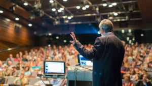 A event host speaking in front of the audience with 2 monitors on the podium.