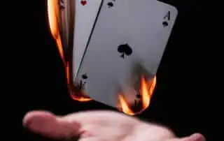 A dramatic scene of four Ace playing cards, engulfed in flames at the edges, held above an open hand against a dark background.