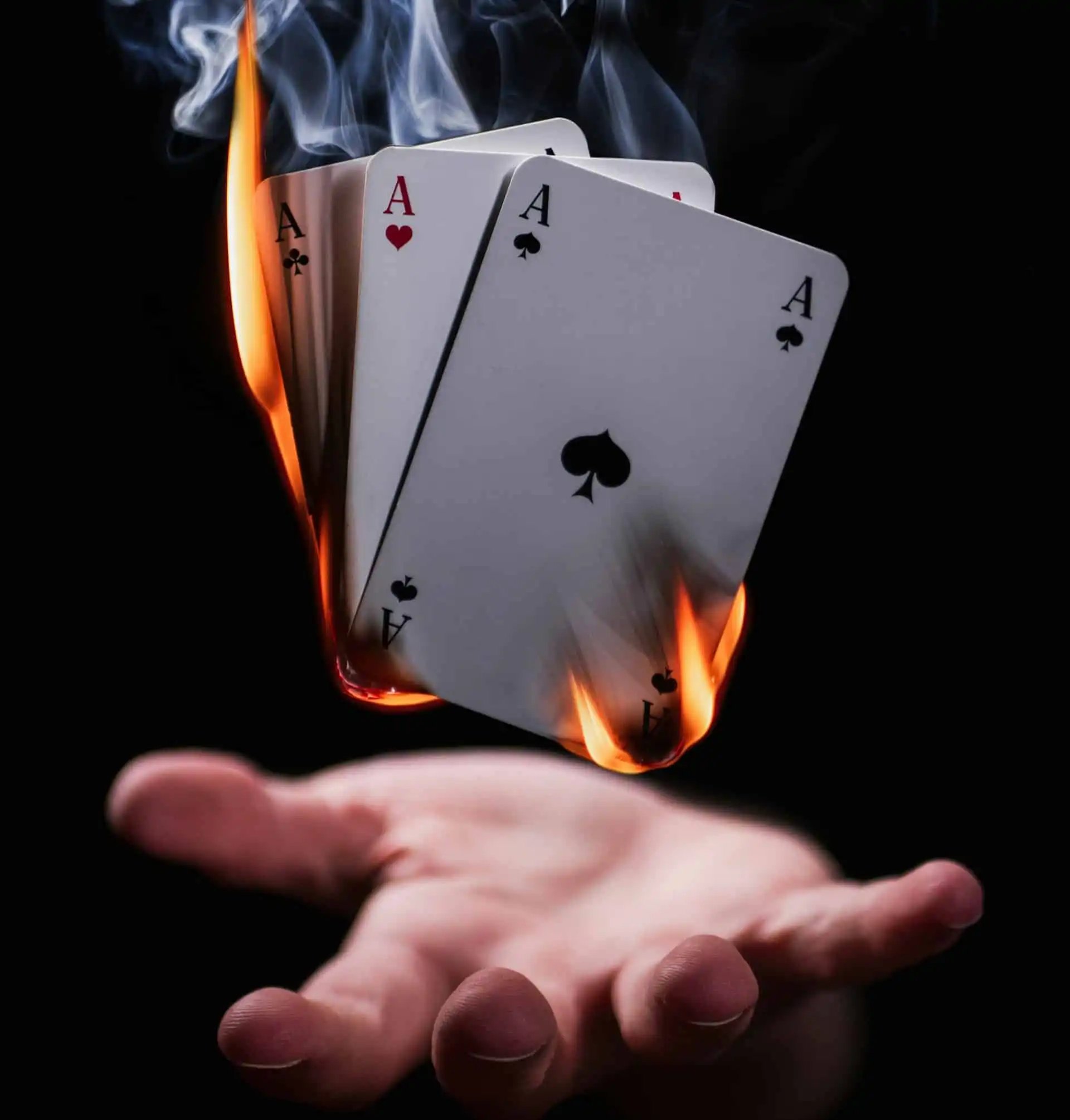 A dramatic scene of four Ace playing cards, engulfed in flames at the edges, held above an open hand against a dark background.