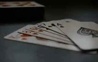 A fan of playing cards featuring the 8 of Hearts, 9 of Hearts, 7 of Spades, and Ace of Spades, laid on a dark surface with a deck of cards in the background