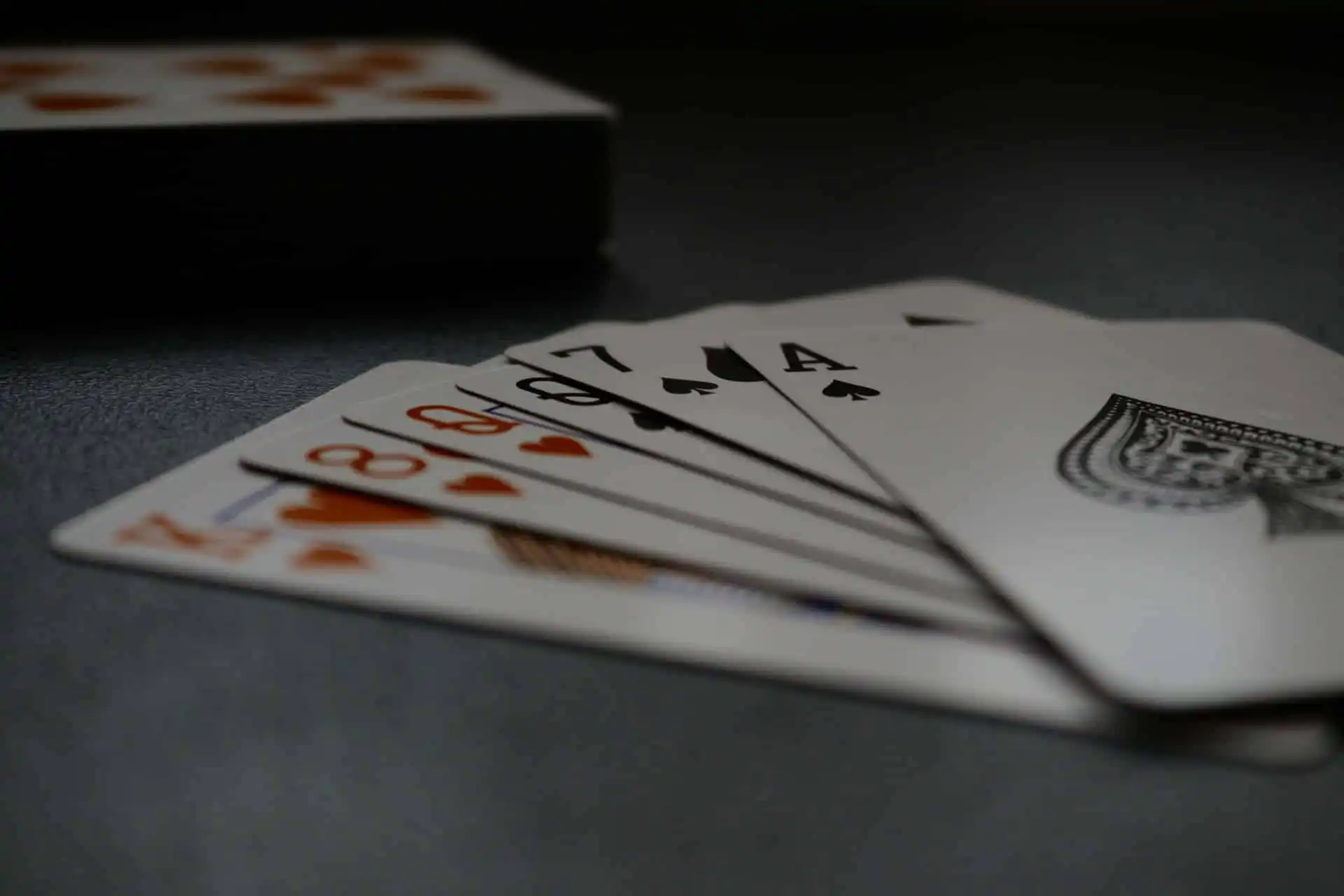 A fan of playing cards used by magicians featuring the 8 of Hearts, 9 of Hearts, 7 of Spades, and Ace of Spades, laid on a dark surface with a deck of cards in the background