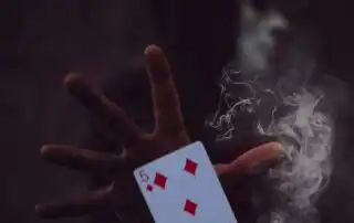 A five of diamonds playing card held by a hand, surrounded by swirling smoke, with a blurred masked figure in the dark background