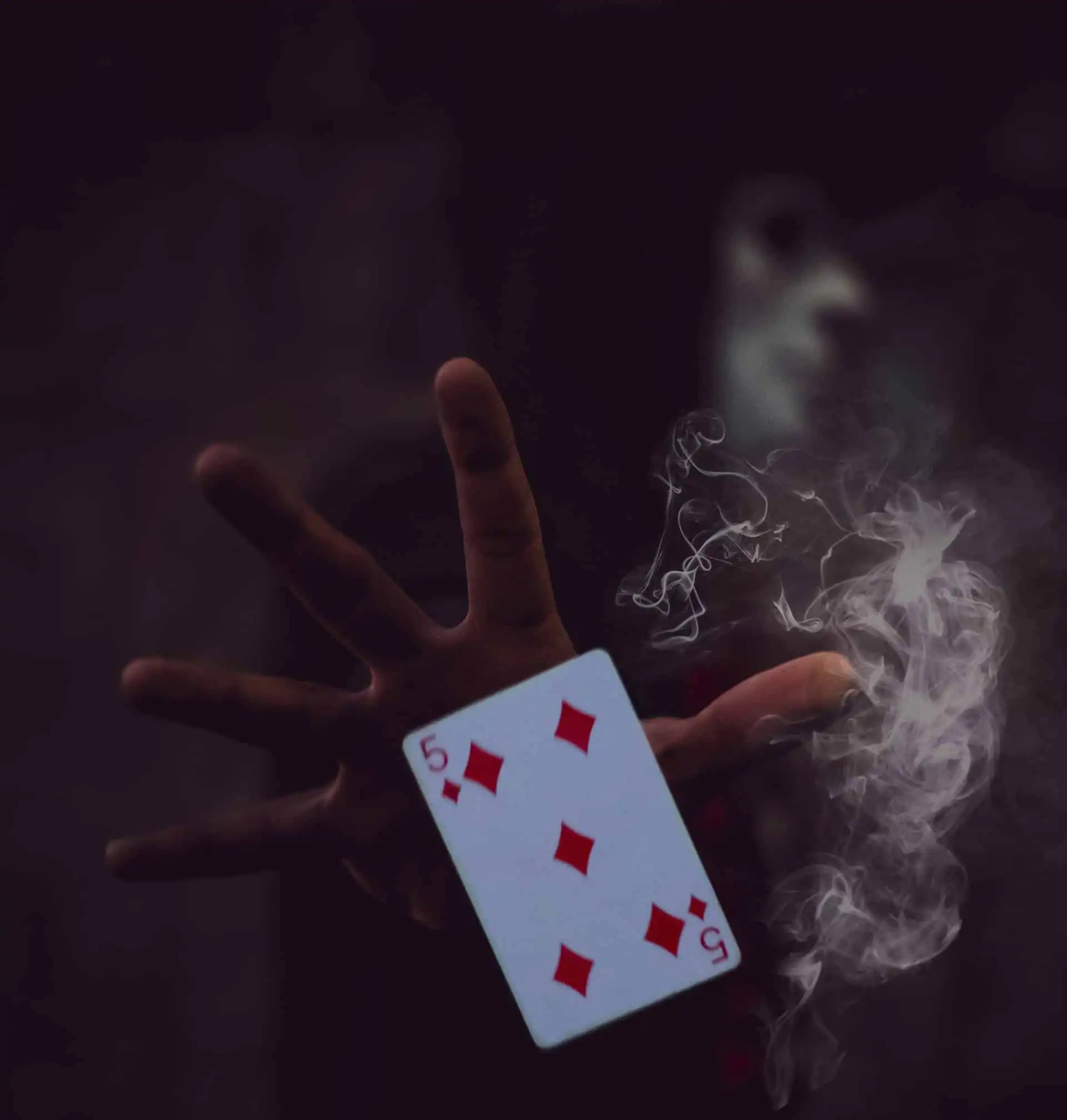 A five of diamonds playing card held by a hand, surrounded by swirling smoke, with a blurred masked figure in the dark background