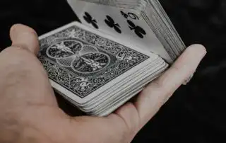 A hand holding a fanned-out deck of playing cards, with the 9 of clubs visible on top, against a dark background.