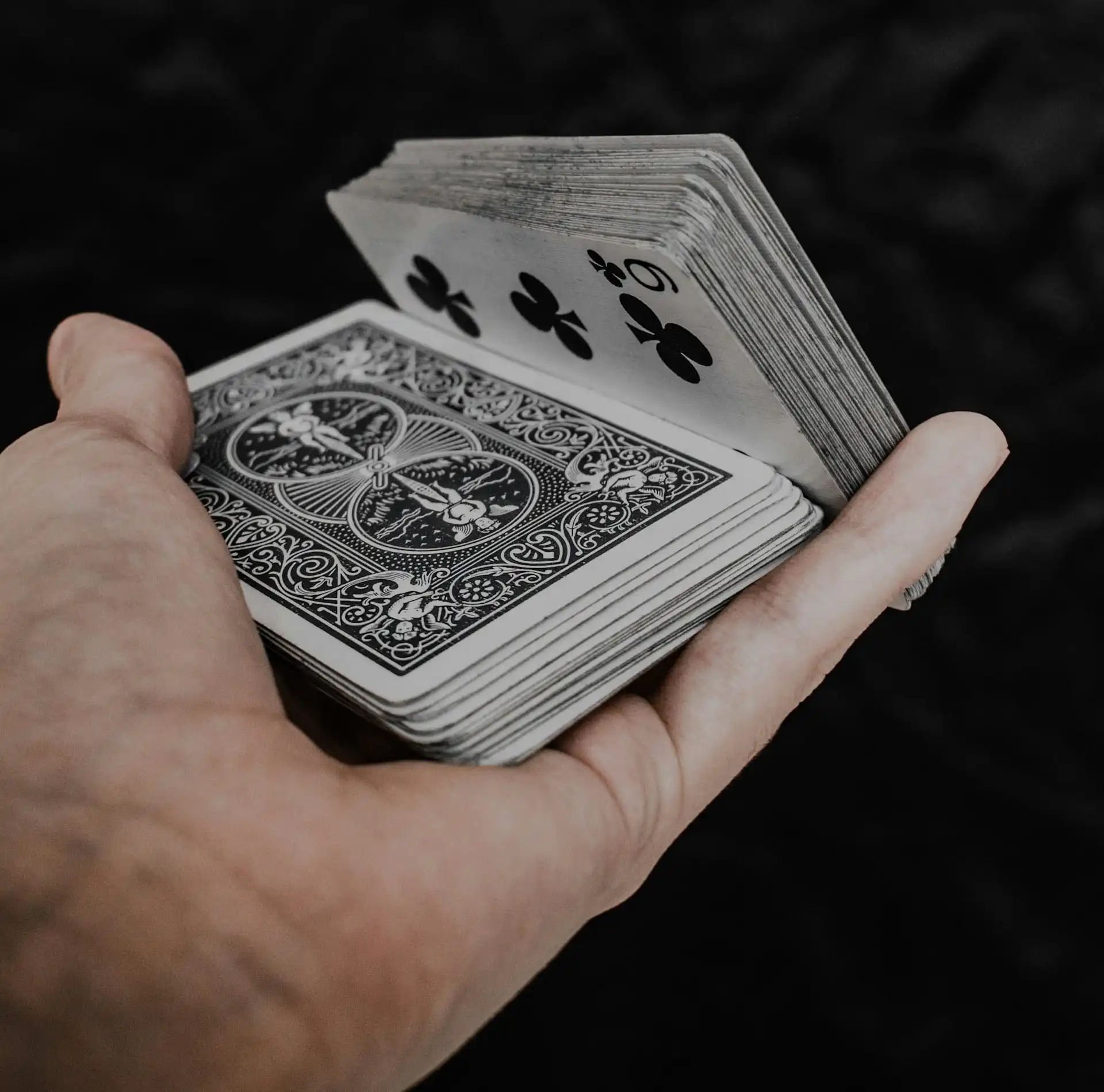 A hand holding a fanned-out deck of playing cards used by magicians, with the 9 of clubs visible on top, against a dark background.