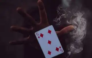 A hand holding the 5 of Diamonds playing card with wisps of smoke around it, set against a dark, mysterious background in a magic shows