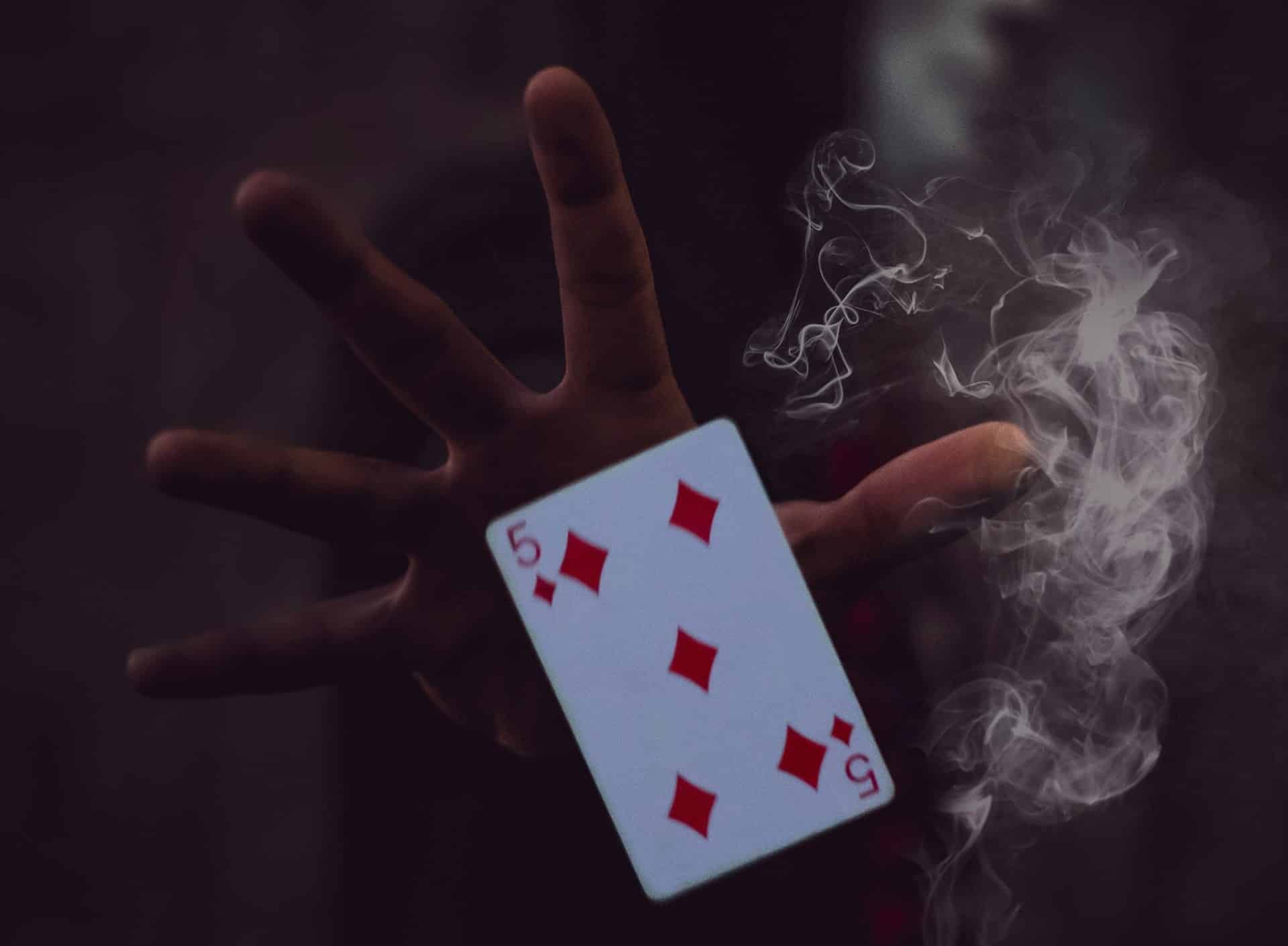 A hand holding the 5 of Diamonds playing card with wisps of smoke around it, set against a dark, mysterious background in a magic shows