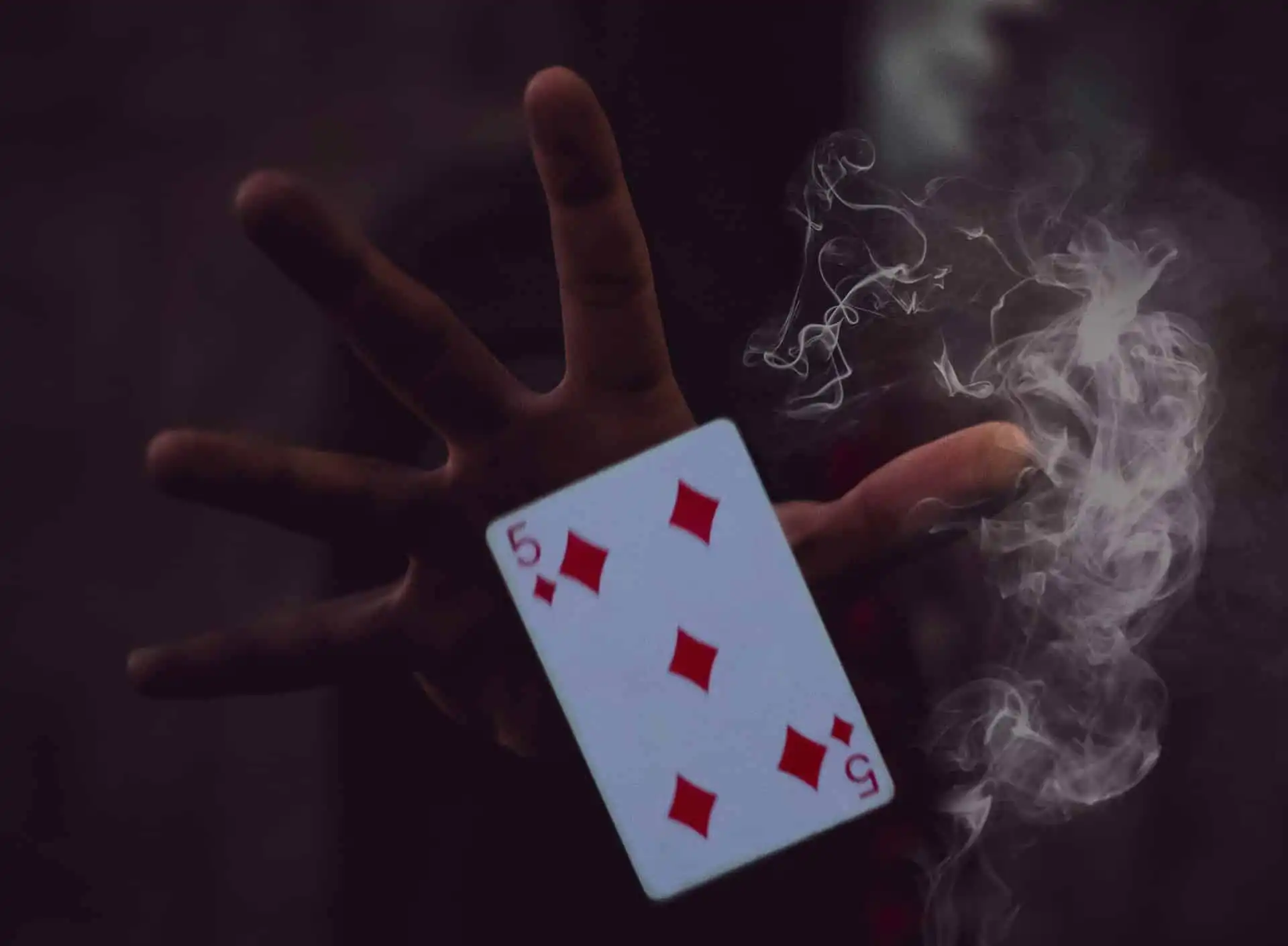 A hand holding the 5 of Diamonds playing card with wisps of smoke around it, set against a dark, mysterious background in a magic shows