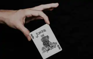 A joker playing card held in a hand against a dark background, featuring the word JOKER, a $ symbol, and an illustration of a jester riding a bicycle