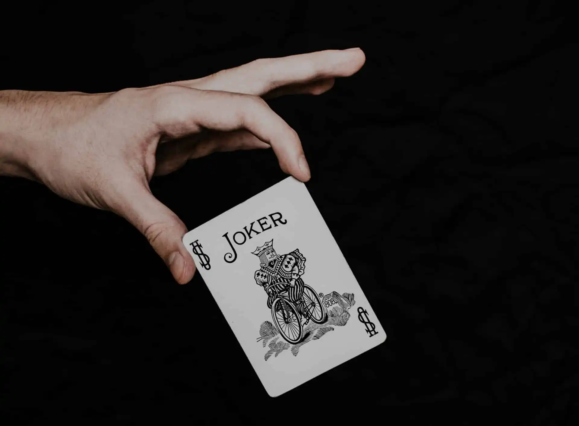 A joker playing card held by a magician in a hand against a dark background, featuring the word JOKER, a $ symbol, and an illustration of a jester riding a bicycle
