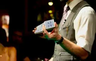 A magician performing a card trick, holding a split deck of blue-backed playing cards in a dimly lit room, wearing a gray vest, white shirt, and purple-striped tie.