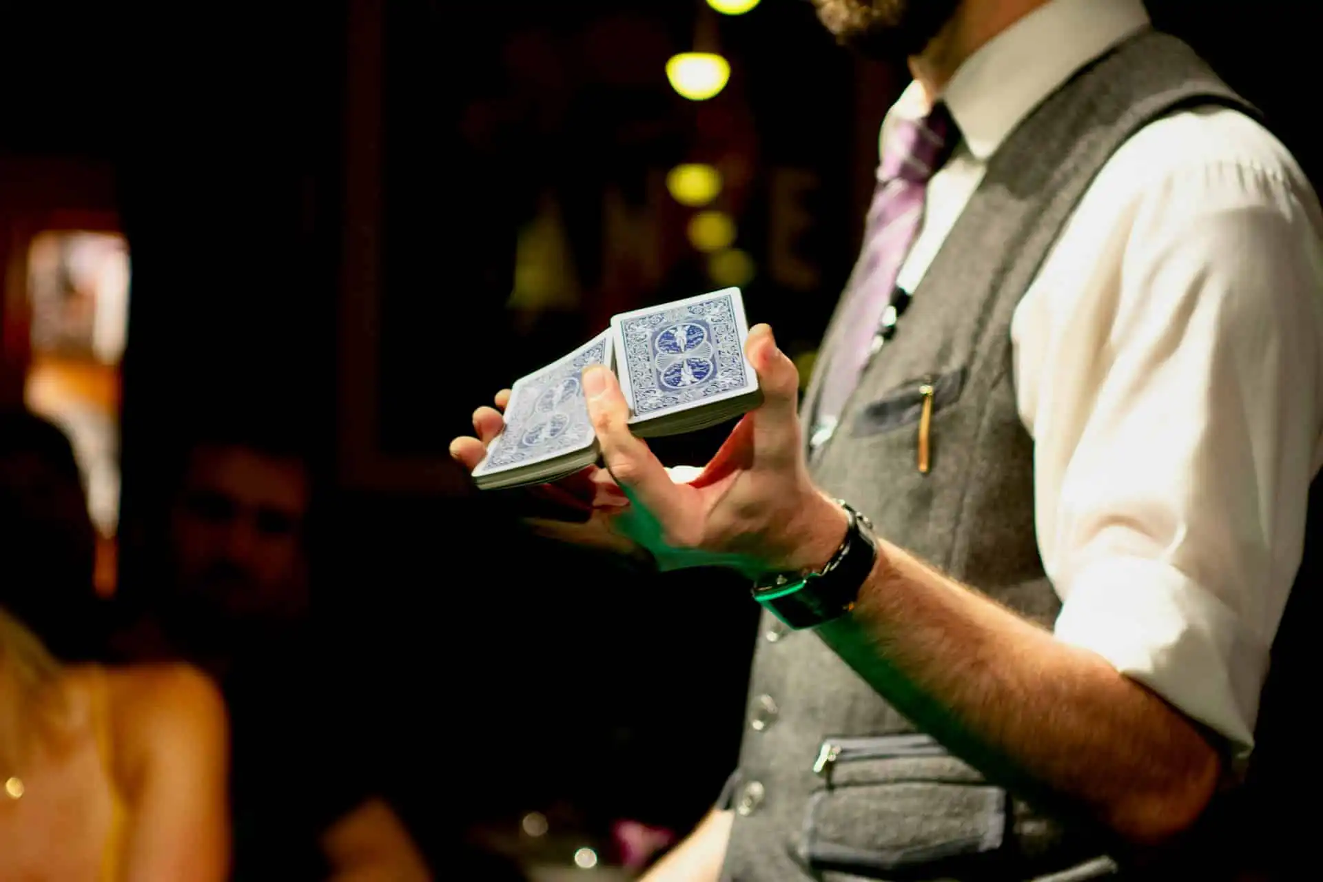 A magician performing a card trick, holding a split deck of blue-backed playing cards in a dimly lit room, wearing a gray vest, white shirt, and purple-striped tie.