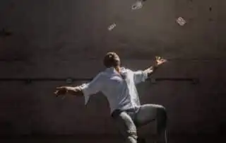 A man in a white shirt and gray pants stands against a textured concrete wall, arms outstretched, with playing cards suspended mid-air above him.