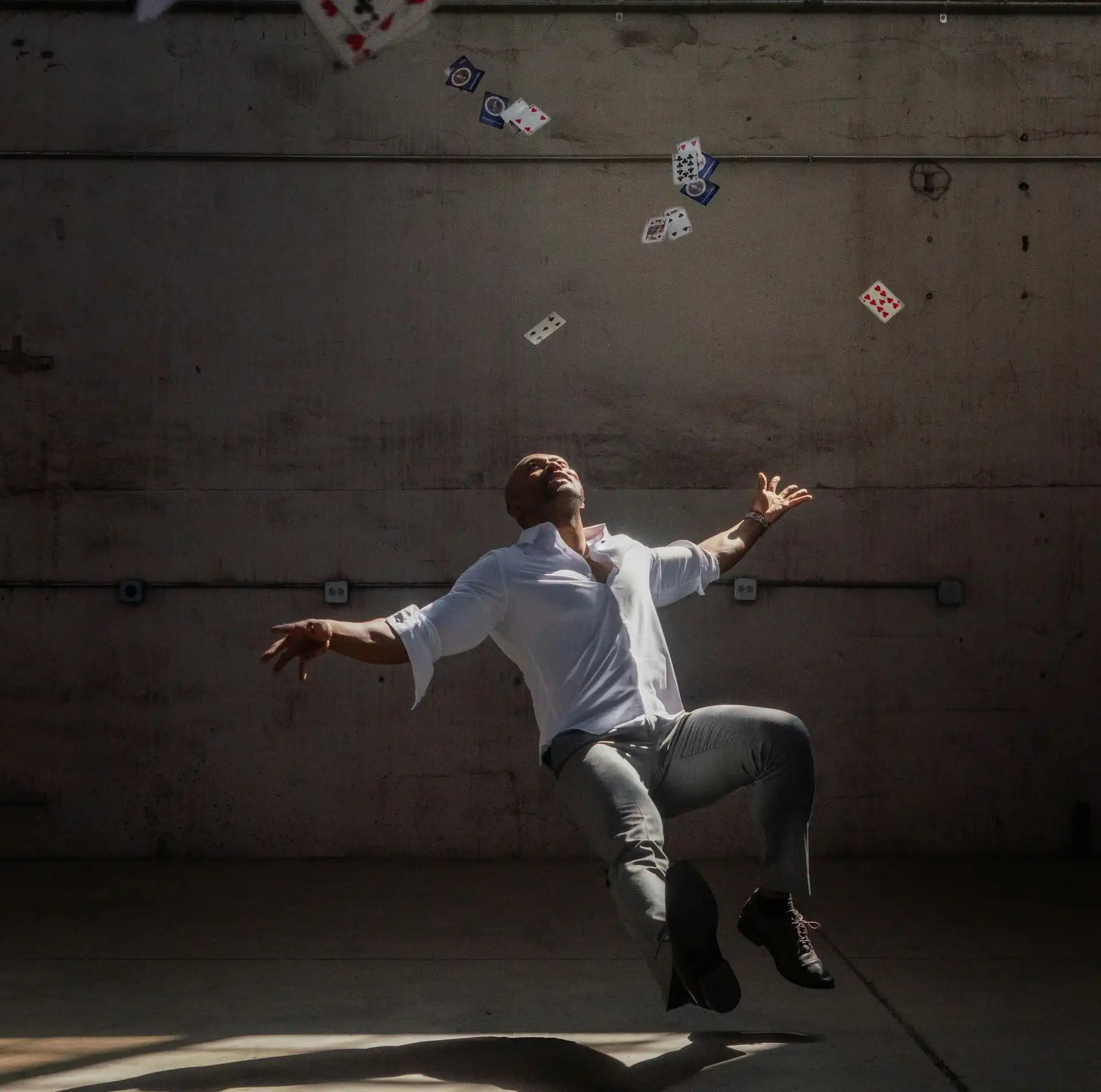 A man in a white shirt and gray pants stands against a textured concrete wall, arms outstretched, with playing cards suspended mid-air above him.