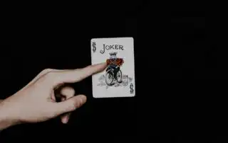 A playing card featuring a joker riding a bicycle, held by a left hand against a black background