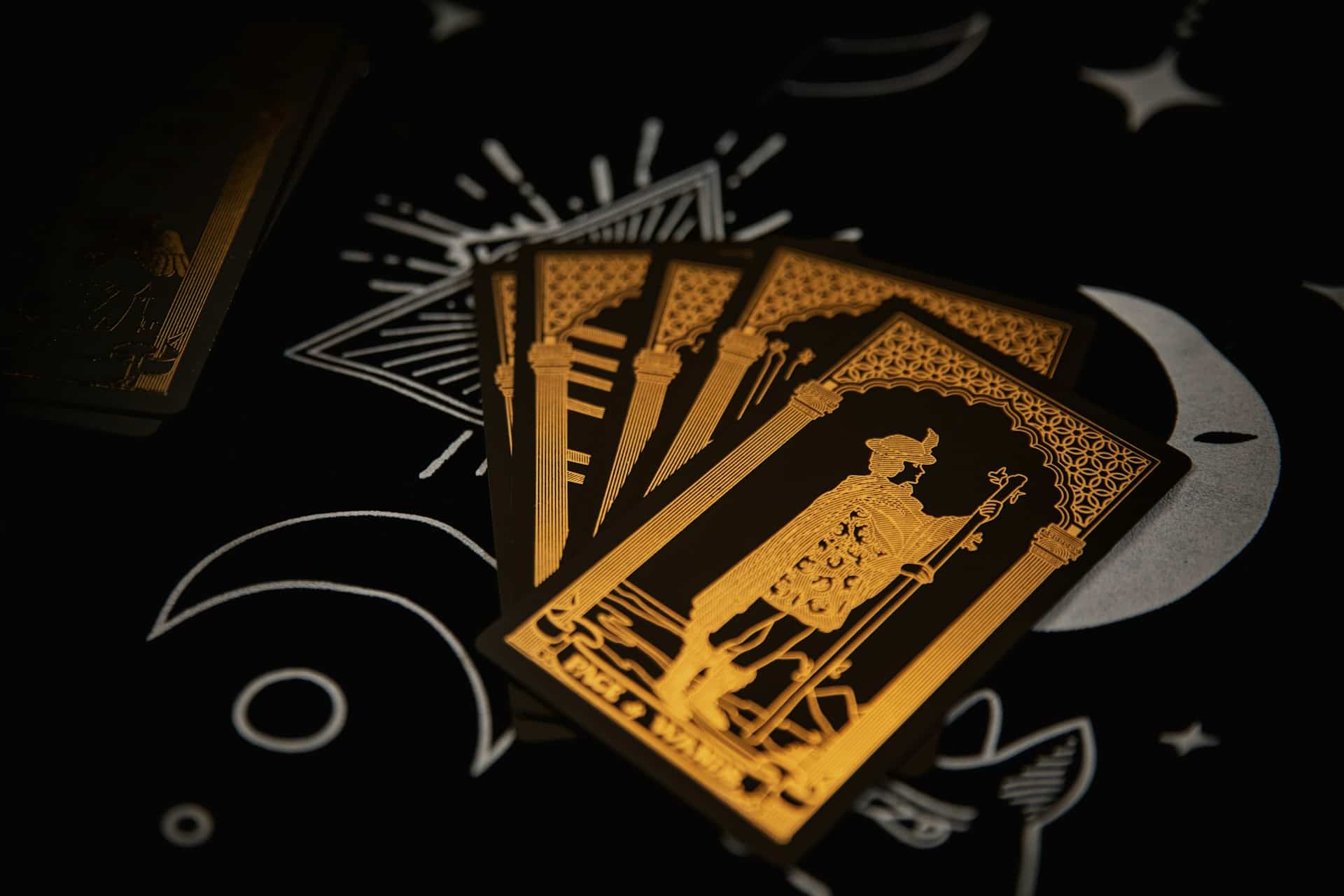 A set of four tarot cards with black and gold designs used by magicians, including the Page of Wands, fanned out on a celestial-themed black background