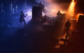 A three-member band performing on stage with vibrant blue and orange lighting, surrounded by fog