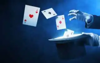 A magical scene with four aces (hearts, clubs, diamonds, spades) floating above a black mentalist hat, with white-gloved hands gesturing against a smoky blue background