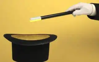 A mentalist gloved hand holding a black magic wand with a white tip, pointing towards a black top hat with a golden interior, set against a yellow background