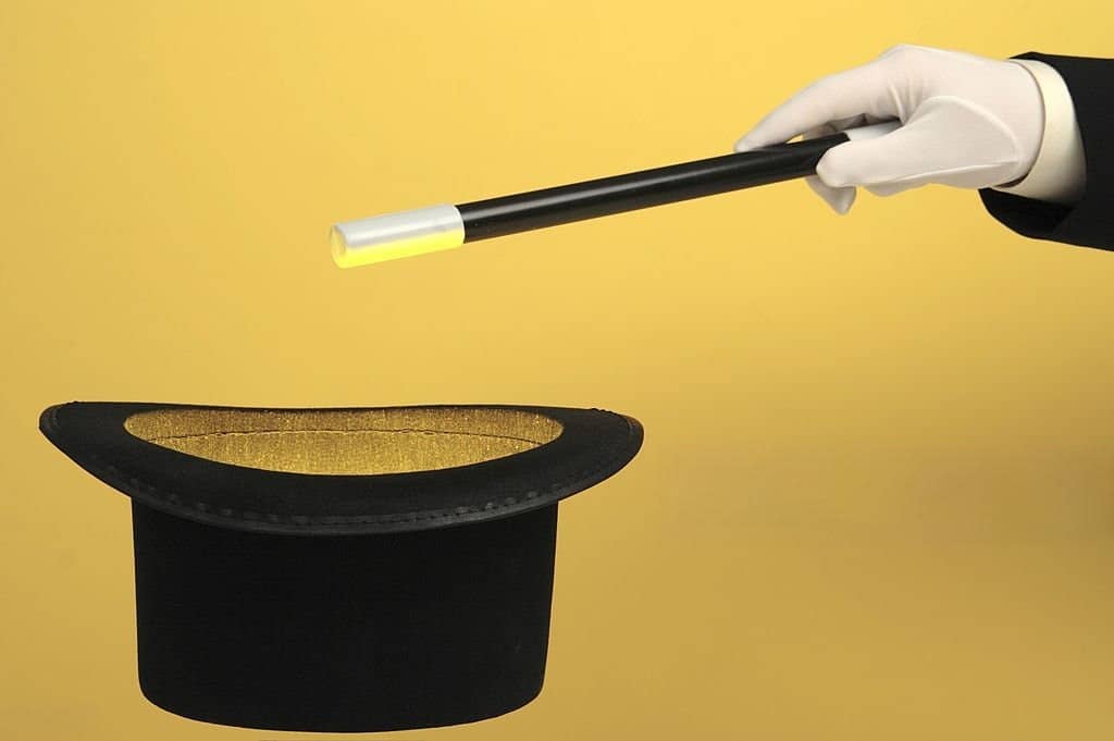 A mentalist gloved hand holding a black magic wand with a white tip, pointing towards a black top hat with a golden interior, set against a yellow background