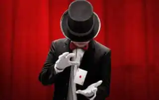 A mentalist in a black suit, red bow tie, and top hat performing a card trick with the Ace of Diamonds floating between their hands, set against a red curtain backdrop
