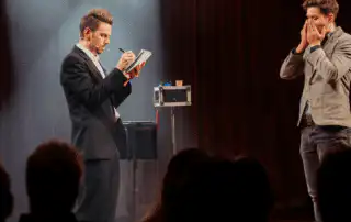 A mentalist on stage during a live show, in a black suit writing on a notepad with a microphone headset