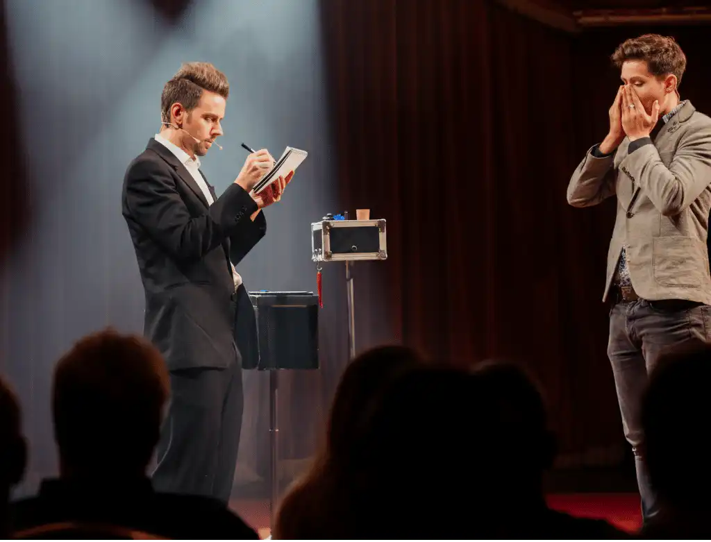 A mentalist on stage during a live show, in a black suit writing on a notepad with a microphone headset