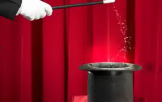 A mentalists gloved hand holding a wand, pointing at a black top hat on a red surface, with golden sparkles and red stage curtains in the background