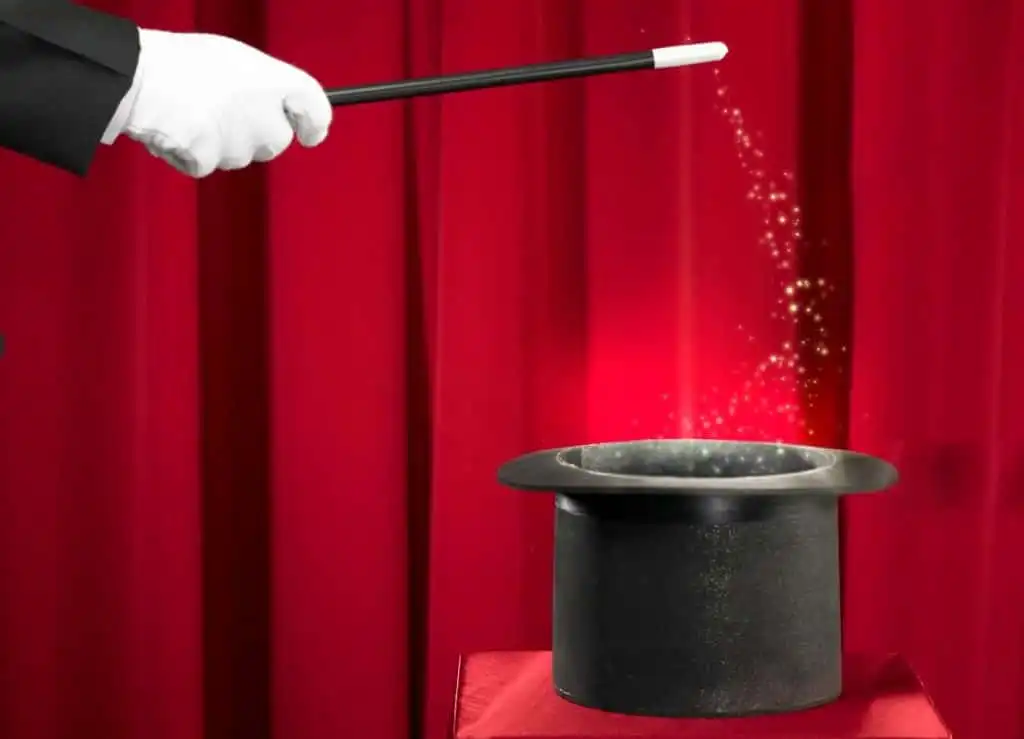 A mentalists gloved hand holding a wand, pointing at a black top hat on a red surface, with golden sparkles and red stage curtains in the background