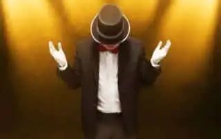 A mentalists in a black suit, red bow tie, and top hat stands under spotlights on a golden textured background, arms slightly raised
