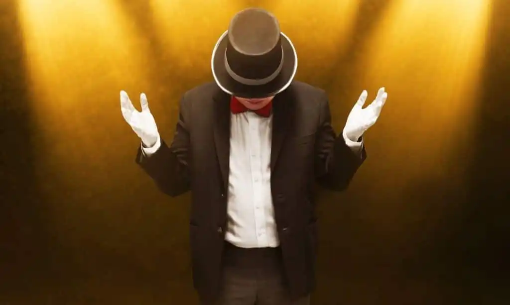 A mentalists in a black suit, red bow tie, and top hat stands under spotlights on a golden textured background, arms slightly raised