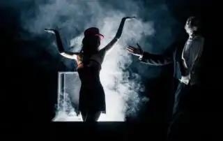Two performers in a theatrical magic act, with one emerging dramatically from a smoke-filled box wearing a red dress and top hat, while the other, in formal attire, gestures in presentation