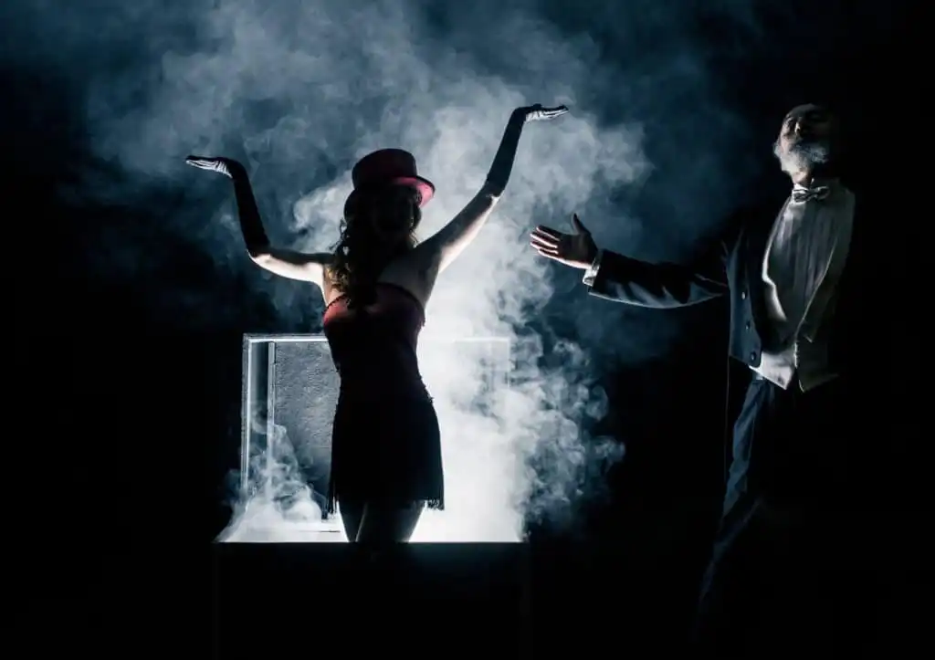 Two mentalists in a theatrical magic act, with one emerging dramatically from a smoke-filled box wearing a red dress and top hat, while the other, in formal attire, gestures in presentation