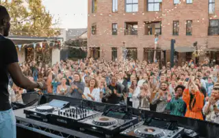 A DJ performing on a raised stage at a lively outdoor team-building event. The DJ is surrounded by a large, diverse crowd is in the background, clapping and enjoying the music