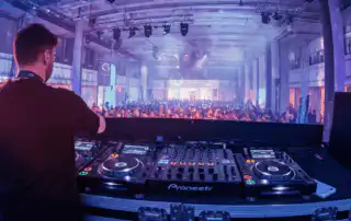 A corporate event with a DJ on stage operating professional equipment, facing a large, energetic crowd while playing his playlist