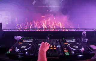 An energetic DJ performance at a live event, featuring professional Pioneer DJ equipment, vibrant purple and pink stage lighting, and a lively crowd with hands raised in the air