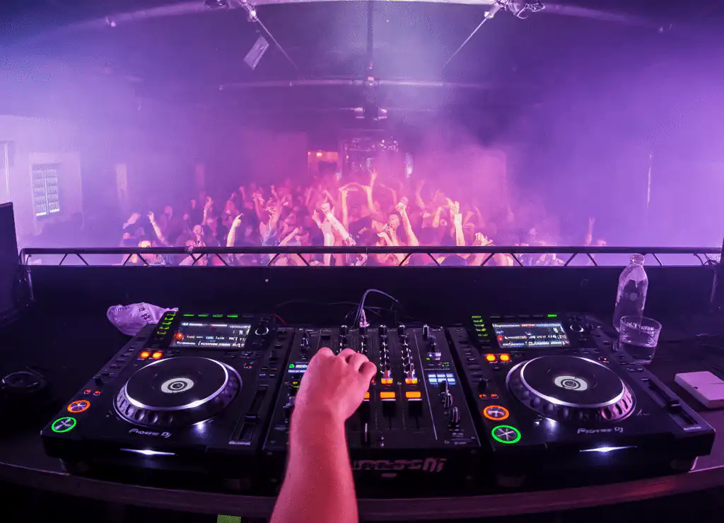 An energetic DJ performance playing different song types at a live event, featuring professional Pioneer DJ equipment, vibrant purple and pink stage lighting, and a lively crowd with hands raised in the air
