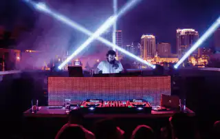 A DJ performing at a rooftop club at night, illuminated by dramatic light beams and a colorful LED screen, with a city skyline backdrop