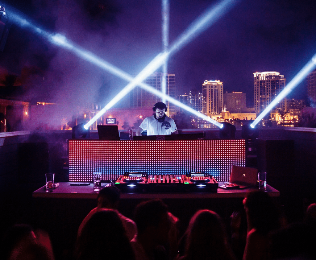A DJ performing at a rooftop club at night, illuminated by dramatic light beams and a colorful LED screen, with a city skyline backdrop