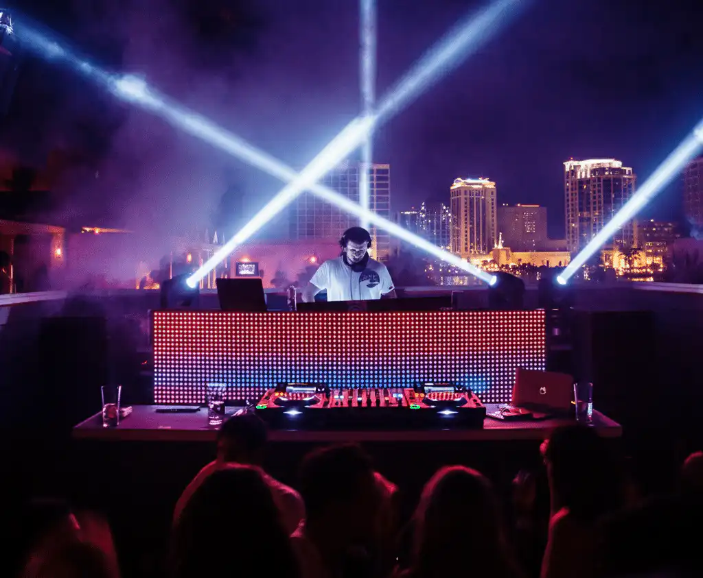 A DJ performing at a rooftop club at night, illuminated by dramatic light beams and a colorful LED screen, with a city skyline backdrop