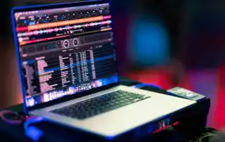A laptop showing a DJ playlists is placed on a flat surface, displaying colorful waveforms and a track list on its screen