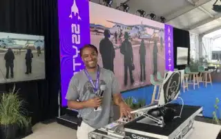 Corporate DJ Will Gill stands at a DJ booth with a microphone, surrounded by a modern event setting.